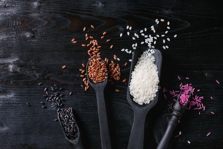 Variety assortment of raw uncooked colorful rice white, black, brown, pink in black spoons and scoop over burnt wooden background. Top view with spaceの写真素材