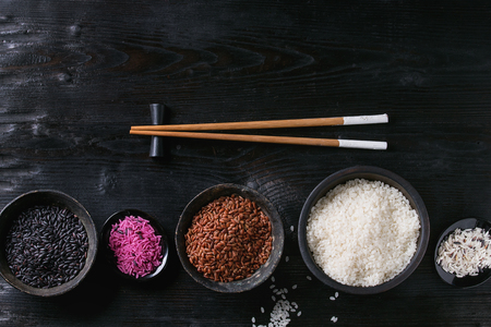 Variety assortment of raw uncooked colorful rice white, black, brown, pink in black bowls over burnt wooden background with chopsticks. Top view with spaceの写真素材