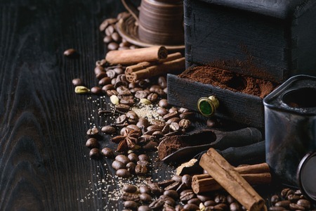 Black roasted coffee beans and grind with spices cinnamon, anise, cardamom, clove and brown sugar. With black vintage coffee grinder, scoops and terracota cup over wood burnt background.の写真素材