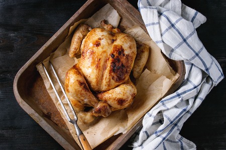 Grilled baked whole organic chicken on backing paper in old oven tray with white kitchen towel and meat fork over black burnt wooden background. Top view with space.の写真素材