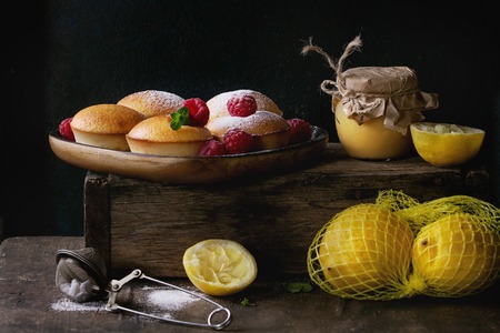 Homemade lemon muffin cupcakes with fresh raspberries, sugar powder, mint, served with jar of lemon curd and lemons on wooden box over dark background. Dark rustic style.の写真素材