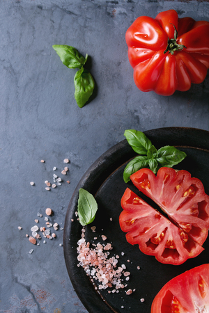 Whole and sliced organic tomatoes Coeur De Boeuf. Beefsteak tomato with pink salt and basil on black wooden chopping board over blue gray metal texture background. Top view with space.の写真素材