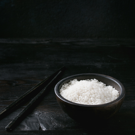 White sushi raw uncooked rice in black bowl with chopsticks over burnt wooden background. Copy space. Square imageの写真素材