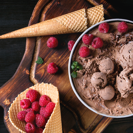 Bowl of homemade chocolate ice cream with fresh raspberries, mint and waffle cone on wood serving board over black burnt wooden background. Top view. Square imageの写真素材