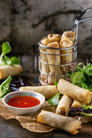 Fried spring rolls with red pepper sauce, served on paper and in fry basket with fresh green salad over old metal texture background. Top view. Asian foodの写真素材