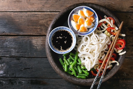 Ingredients for cooking stir fry udon noodles, green beans, sliced paprika, boiled eggs, soy sauce with sesame seeds in traditional bowls with wooden chopsticks over old wooden background. Flat layの写真素材