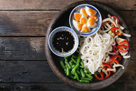 Ingredients for cooking stir fry udon noodles, green beans, sliced paprika, boiled eggs, soy sauce with sesame seeds in traditional bowls in terracotta tray over old wooden background. Flat layの写真素材