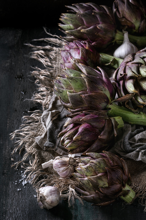 Uncooked whole organic wet purple artichokes with garlic on textile sackcloth over dark wooden background. Rustic style.の写真素材