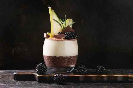 Dessert breakfast layered chia seeds, chocolate pudding, rice porridge in glass decorated by fresh blackberries, pear, cocoa powder. Stand with spoon on serving board over dark texture background.の写真素材