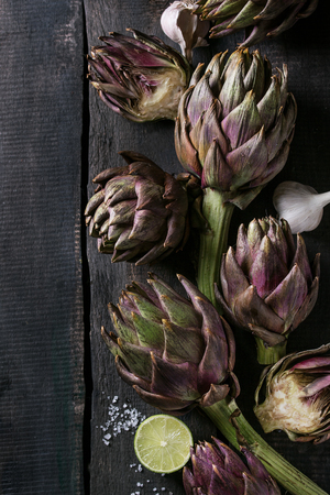 Uncooked whole and sliced organic wet purple artichokes with salt, lime and garlic over dark wooden background. Rustic style. Top view with spaceの写真素材