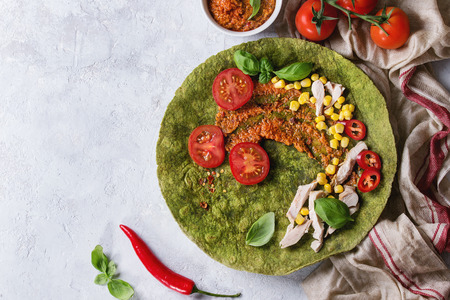 Green spinach matcha tortilla with ingredients for filling ready to wrap. Sweet corn, tomatoes, chili pepper, chicken meat, basil served over gray texture background and textile. Flat layの写真素材