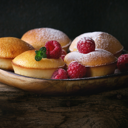 Homemade lemon muffin cupcakes with fresh raspberries, sugar powder, mint, served in square wood plate on wooden table over dark background. Dark rustic style. Close up. Square imageの写真素材