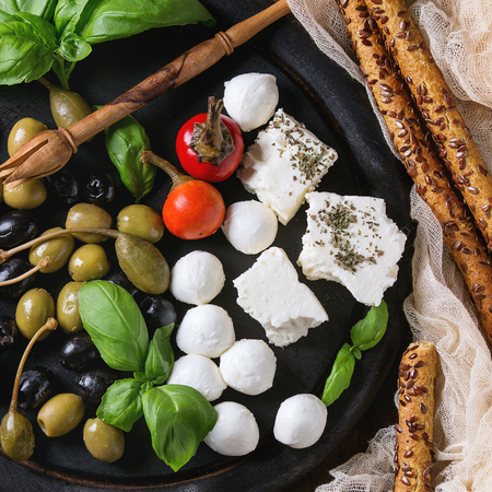Mediterranean appetizer antipasti board with green black olives, feta cheese, mozzarella, capers, pepper, basil with grissini bread sticks over old wooden background. Top view. Square imageの写真素材