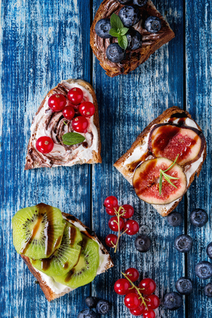 Variety of dessert sandwiches with berries and cream cheese and chocolate swirl. Red currant, blueberries, sliced kiwi, figs over blue wooden plank background. Flat lay, summer appetizer conceptの写真素材