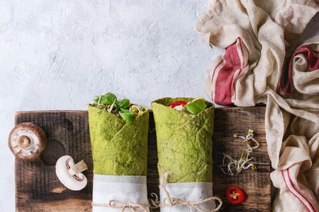 Green spinach matcha tortillas wrapped in paper with ingredients above. Sweet corn, avocado, basil, sprouts, mushrooms served on wood board over gray texture background and textile. Flat layの写真素材