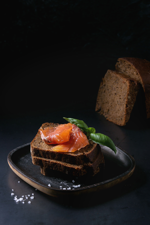 Stack of sliced homemade rye bread with smoked salmon, sea salt and fresh basil on wooden plate over dark black background.の写真素材