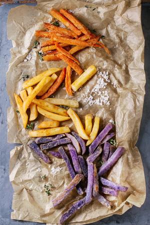 Variety of french fries traditional potatoes, purple potato, carrot served with salt, thyme on baking paper over gray metal background. Top view with space. Homemade fast foodの写真素材