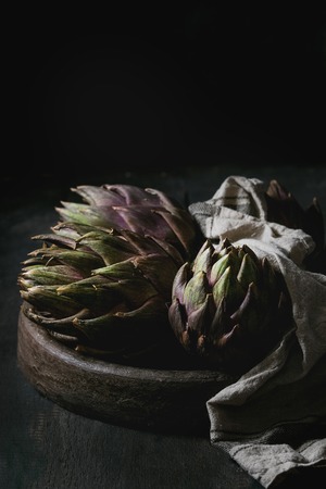Uncooked whole organic wet purple artichokes in terracotta tray with textile over dark wooden background. Rustic style.の写真素材
