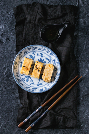 Japanese Rolled Omelet Tamagoyaki sliced with black seasame seeds and soy sauce, served in blue white ornate ceramic plate with chopsticks, textile over black stone background. Top view with spaceの写真素材