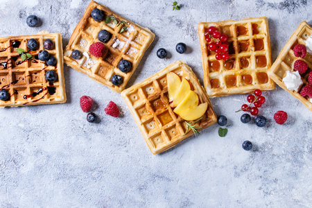 Homemade square belgian waffles with fresh ripe berries blueberry, raspberry, red currant, peach served with caramel, balsamic sauce, honey, whipped cream over gray background. Top view with spaceの写真素材