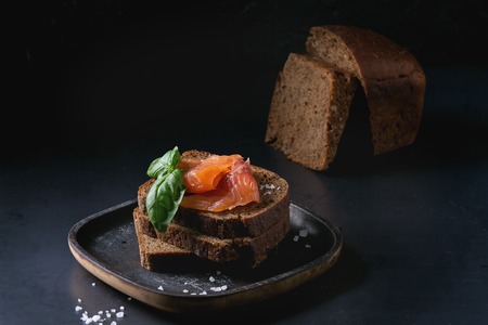 Stack of sliced homemade rye bread with smoked salmon, sea salt and fresh basil on wooden plate over dark black background.の写真素材