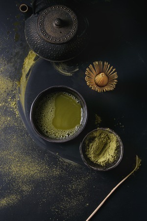 Green tea matcha powder and hot drink in black bowls standing with iron teapot, bamboo traditional tools spoon and whisk over dark metal background. Top view with spaceの写真素材