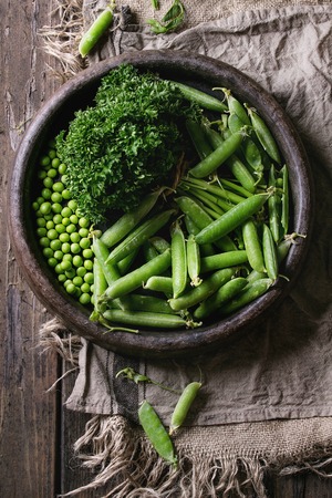 Young organic green pea pods and peas in terracotta tray and bundle of parsley over old dark wooden planks with sackcloth textile background. Top view with space. Harvest, healthy eating.の写真素材