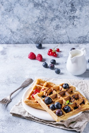 Breakfast with homemade square belgian waffles with fresh ripe berries blueberry, raspberry, red currant, balsamic sauce on white plate with jug of milk, textile napkin over gray texture background.の写真素材