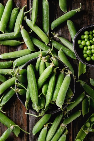 Young organic green pea pods and peas in bowls over old dark wooden planks background. Top view with space.の写真素材