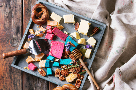Variety of colorful holland cheese traditional soft, old, pink basil, blue lavender served with pecan nuts, honey, lavender flowers, pretzels bread on tray over wooden planks background. Flat layの写真素材
