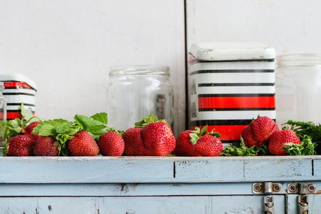 Fresh ripe garden strawberries and melissa herbs with empty glass and vintage metal jars for jam on blue white wooden kitchen table. Rustic style, day light, copy spaceの写真素材