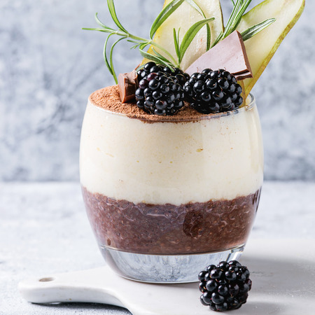 Dessert breakfast layered chia seeds, chocolate pudding, rice porridge in glass decorated by fresh blackberries, sliced pear, cocoa. Stand on white serving board over gray background. Square imageの写真素材