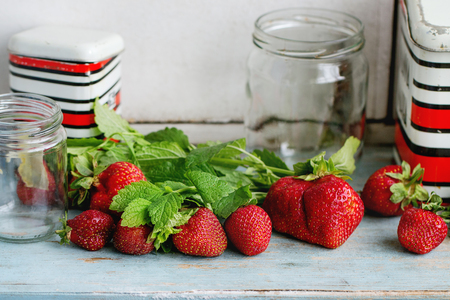 Fresh ripe garden strawberries and melissa herbs with empty glass and vintage metal jars for jam on blue white wooden kitchen table. Rustic style, day light, copy spaceの写真素材