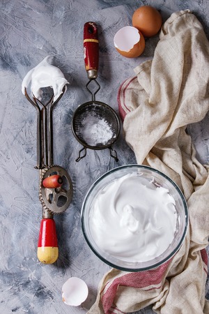 Ingredients and utensils for cooking meringue. Whipped egg whites, sugar powder, broken eggs, vintage sieve and hand mixer on linen towel over gray texture background. Top view. Baking dessert conceptの写真素材