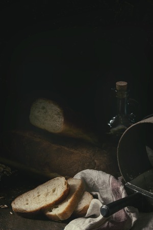 Homemade white wheat bread whole and slice served with bottle of olive oil and wheat grain seeds on white linen towel over dark wooden kitchen table. Rustic style.の写真素材