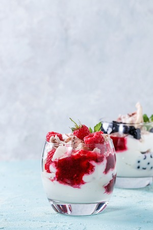 Traditional summer dessert Eton Mess. Broken meringue with whipped cream, berry jam, fresh blueberries and raspberries in two glasses, decorated with mint leaves over light blue concrete background.の写真素材