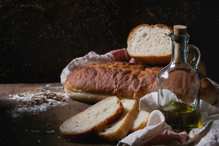 Homemade white wheat bread whole and slice served with bottle of olive oil and wheat grain seeds on white linen towel over dark wooden kitchen table. Rustic style.の写真素材