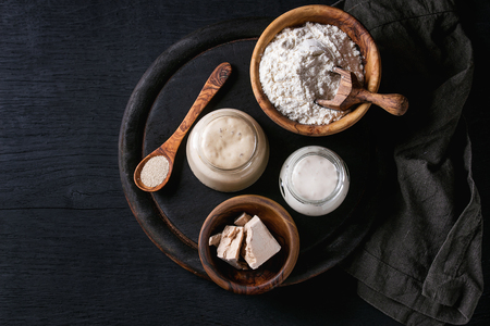 Rye and wheat sourdough in glass jars, fresh and instant yeast, olive wood bowl of flour for baking homemade bread. With spoon, serving board, textile over black burned wooden background. Top viewの写真素材