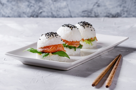 Mini rice sushi burgers with smoked salmon, green salad and sauces, black sesame served on white square plate with wooden chopsticks over gray concrete background. Modern healthy foodの写真素材