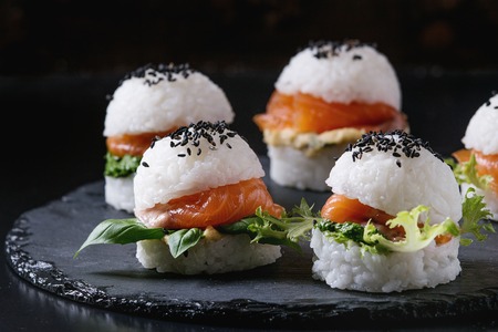 Mini rice sushi burgers with smoked salmon, green salad and sauces, black sesame served on slate stone board over black background. Modern healthy food. Close upの写真素材