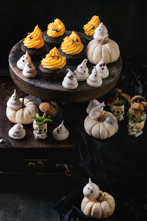 Halloween decorated sweet dessert table black cupcake with orange cream, white meringue ghosts with chocolate eyes, decor skulls and pumpkin over black tablecloth.の写真素材