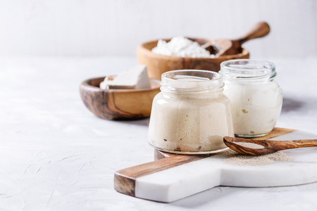 Rye and wheat sourdough in glass jars, fresh and instant yeast, olive wood bowl of flour for baking homemade bread. With spoon, serving board over gray concrete background.の写真素材