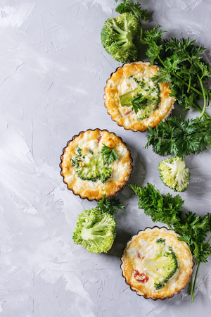 Baked homemade vegetable broccoli quiche pie in mini metal forms served with fresh greens on gray concrete background. Flat lay with copy space. Ready for eatの写真素材