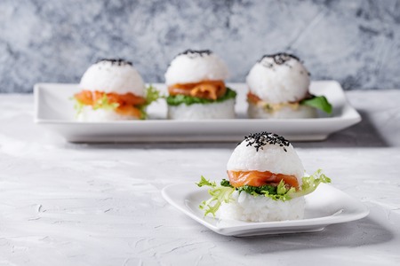 Mini rice sushi burgers with smoked salmon, green salad and sauces, black sesame served on white square plate over gray concrete background. Modern healthy foodの写真素材