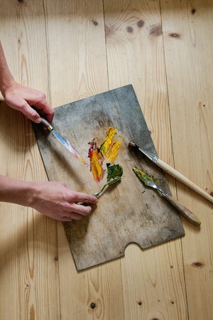 Wooden palette with oil paints yellow red, brushes and palette knife over wooden floor. With female hands in action. Top view with space. Art concept. Atmospheric day lightの写真素材