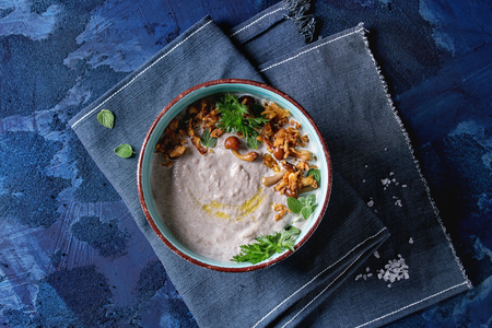 Mushroom cream soup in ceramic bowl served with forest mushrooms, greens, fried onion on blue textile napkin over dark blue texture concrete background. Top view with spaceの写真素材
