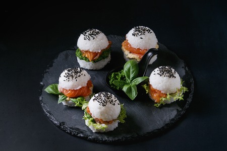 Mini rice sushi burgers with smoked salmon, green salad and sauces, black sesame served on slate stone board over black background. Modern healthy foodの写真素材