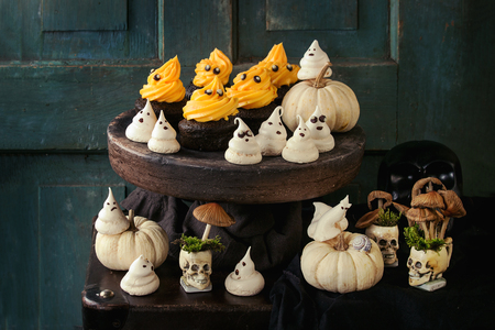 Halloween decorated sweet dessert table black cupcake with orange cream, white meringue ghosts with chocolate eyes, decor skulls, mushrooms and pumpkin over black tablecloth.の写真素材