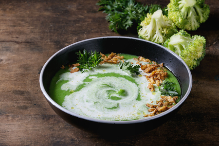 Vegetarian broccoli cream soup served in black bowl with cream, fried onion, fresh parsley and broccoli over old wooden background. Close up. Healthy eating.の写真素材