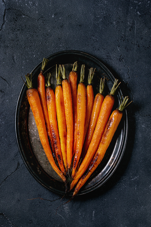 Roasted young whole carrot served on vintage metal tray over black texture background. Top view with spaceの写真素材
